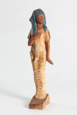 Standing figure of painted plaster with a mummy wrapping dress in tan and a woman emerging from the top with brown skin and a blue head dress.