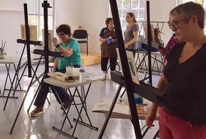 Group of four adults scattered in a large white room, two standing at black metal easels painting on small pieces of paper. There are small grey tables at their sides holding supplies.