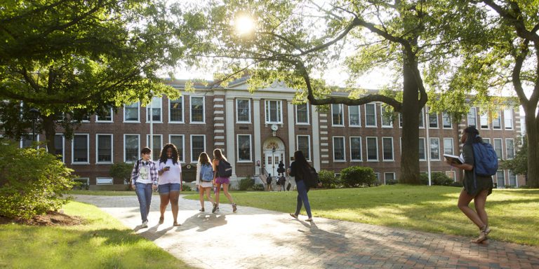 Photograph of a sunny day with green grass and a brick walkway. There are several tall trees on either side. In the background is a long two story brick building with white trim. Throughout are college students walking.