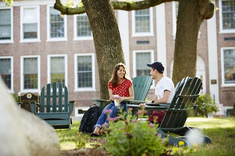 Photograph of a sunny outside with green grass and two large tree trunks. There are four green Adirondack chairs in a semi circle with a woman and man seated in two. In the background is a brick building with white trim.