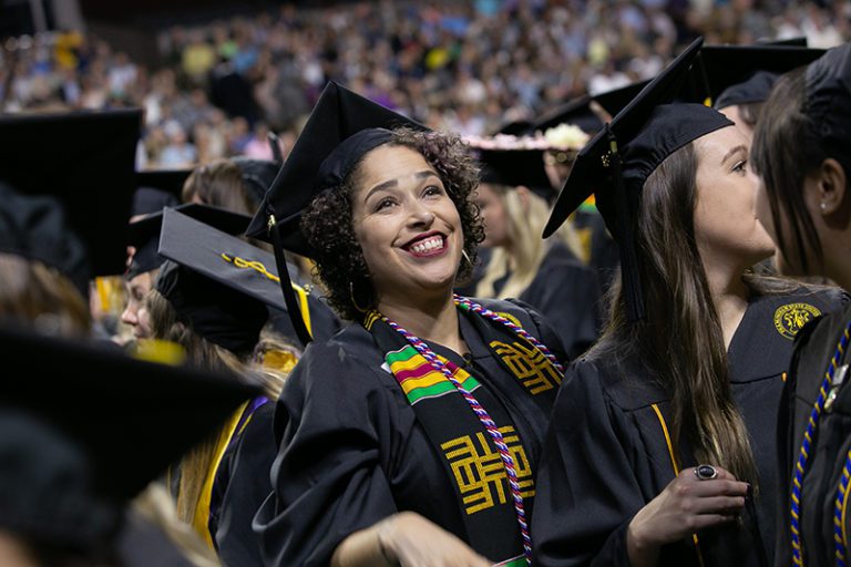 Photograph of a college graduation, zoomed in on a light brown skinned woman with short curly hair in a crowd of other graduates. She is smiling and looking upwards at something off scene.