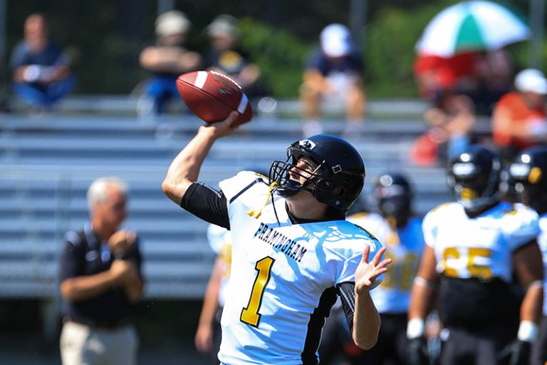 Photograph of a football player in a white jersey and black helmet with a yellow number 1 on his chest. He is in mid action of throwing a football. In the background are blurred stadium seats with a few people milling about.