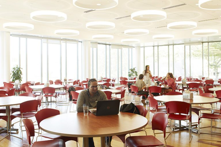 Photograph of an interior room with two walls of windows and circular lights on the ceiling. On the floor are many round wood tables with many red chairs around each table. There is one man at the closest table looking at a lap top and a group of three woman at a father back table.