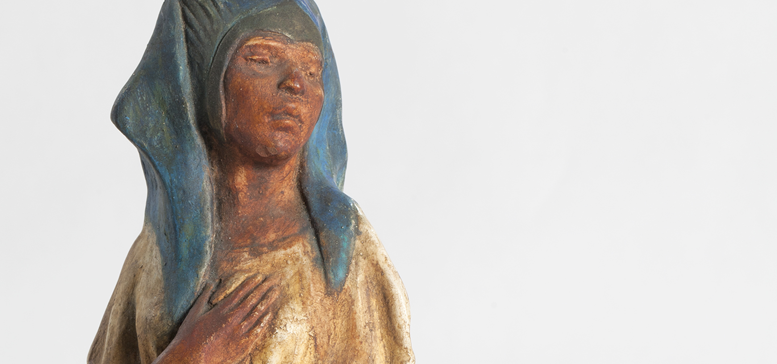 Painted sculpture of a tan skinned woman in a cream dress and blue headpiece, with one hand to chest.