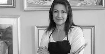 Photograph of a woman with light skin, long brown hair, wearing a white shirt and dark apron, standing in front of a wall filled with framed artworks.