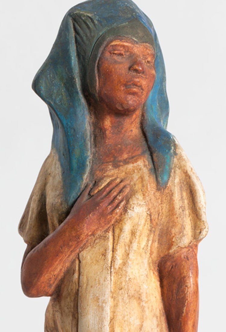 Painted sculpture of a tan skinned woman in a cream dress and blue headpiece, with one hand to chest.