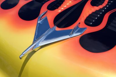 Zoomed in photograph of a hot rod car with red and yellow flames licking a black car body. The focus is on the silver jet plane hood ornament.