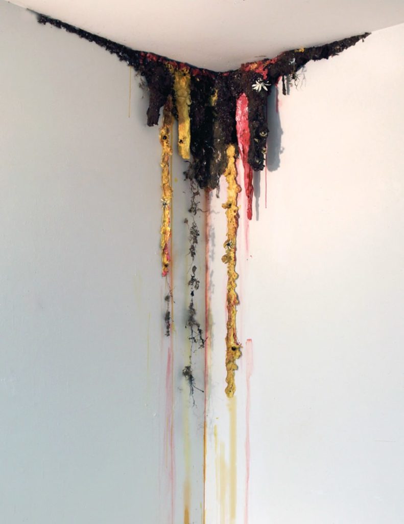Sculpture mounted from a ceiling corner with brown, pink, and yellow oozing protrusions, and dangling yellow and blue pieces of clay and fabric.