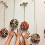Photograph of three small hands on a wall beneath decorative yarn balls on string hangers.