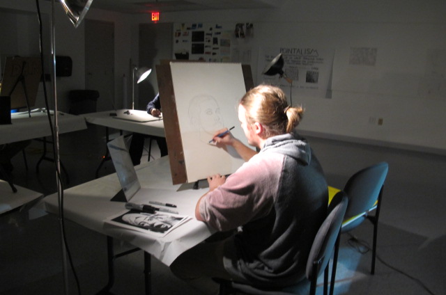 Photograph of a teenaged girl seated at a table with a table easel and a mirror, drawing her reflection in a dimly lit room.