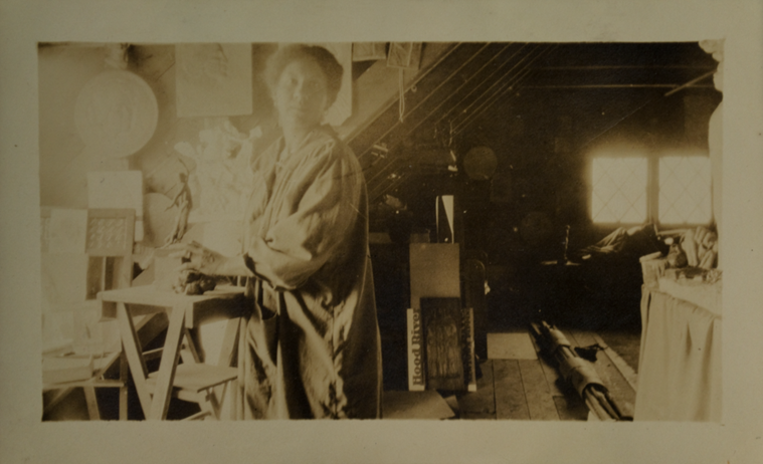 Old photograph of a woman in a baggy work jacket standing in a slanted roofed attic with sculptures all around and a window in the back right.