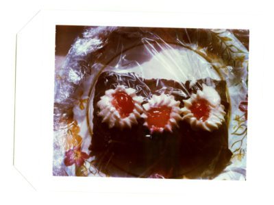 Photograph of a piece of chocolate frosted cake with white and red blob decorations, all wrapped in saran wrap on a patterned paper plate.