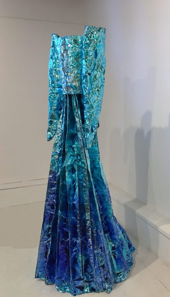 Photograph of a foil dress done in shades of blues with large angular sleeves and a narrow pleated long skirt. The dress is floating in a white room.
