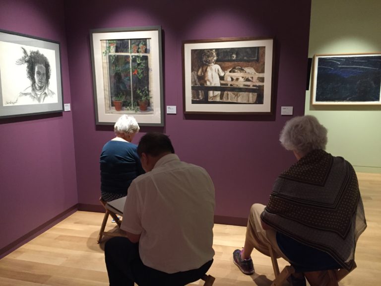Photograph of three older adults seated in small stools drawing in a gallery room with deep eggplant walls, one wall to the far right is light sage green.