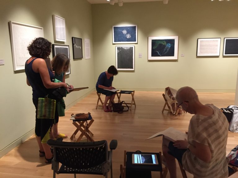 Group of adults, some seated and some standing, around small stools drawing in a gallery room. There are many framed artworks hanging at various levels on the light sage green walls.