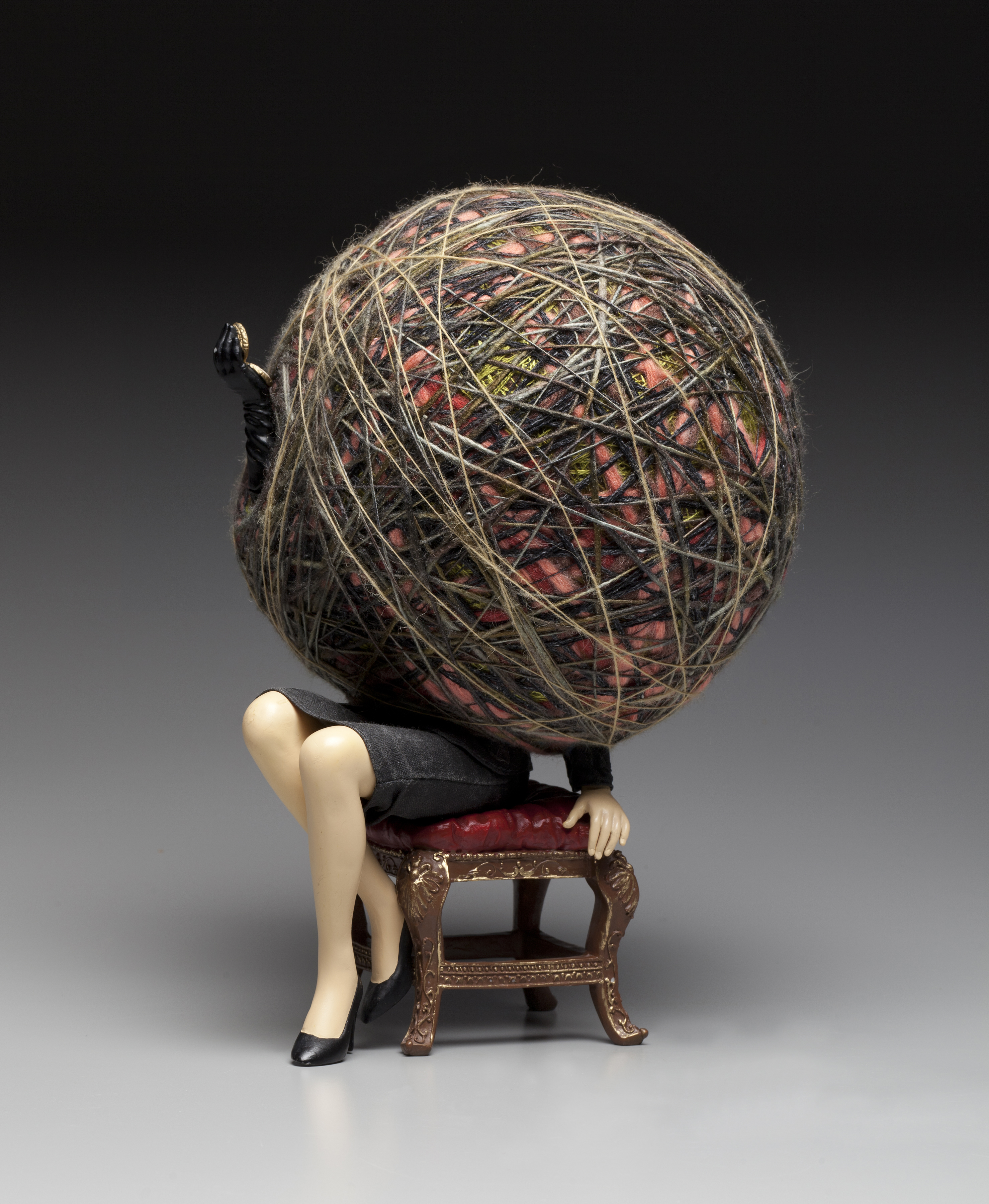 Small sculpture figure of an ornate chair with red cushion where the legs and one arm of a light skinned woman in a pencil skirt is seen. Her entire body is a ball of twine and threads.