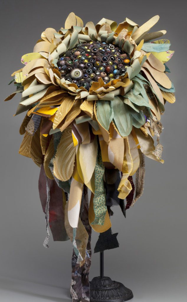 Dramatic hat on a stand, made to look like a sunflower with long petals in soft yellow and light blue-green made of various fabrics, and a black center made of various beads.