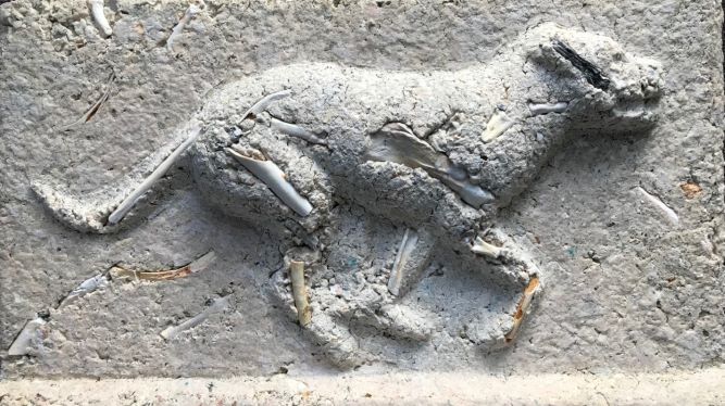 Sculpture with a flat background and a raised dog in the motion of running. There are bones visible throughout the piece.