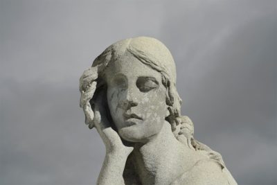 Photograph with a mottled light grey-white sky and a statue in the foreground from the shoulders up of a woman with long hair and hand supporting her head under the chin. The statue is worn, losing details, and a leaf pattern has been imposed over her face.
