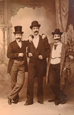 Old photograph of three woman in front of a painted ornate backdrop. All three women are dressed as men, wearing pants, long jackets, and bowler hats with fake mustaches.