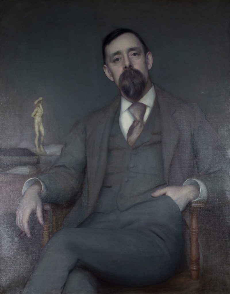 Portrait painting of a cream skinned man, seated, wearing a grey suit. He has short dark hair and a large beard. One hand is in his pants pocket while the other is leaning on a desk next to the chair that has papers and a nude figurine on top.