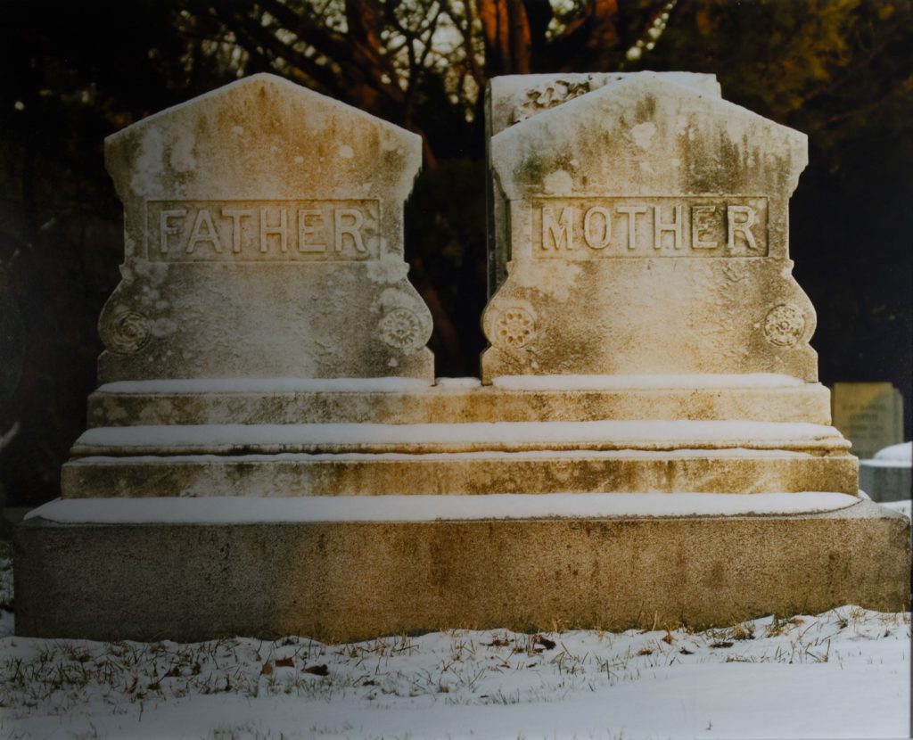 Photograph of a connected pair of yellow-white headstones with 'Father' on the left and 'Mother' on the right. The ground is covered in snow.
