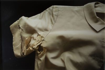 Photograph of a pink-cream blouse, close up focusing on the arm. The short sleeve and armpit are all ragged and shredded. The background is pitch black.
