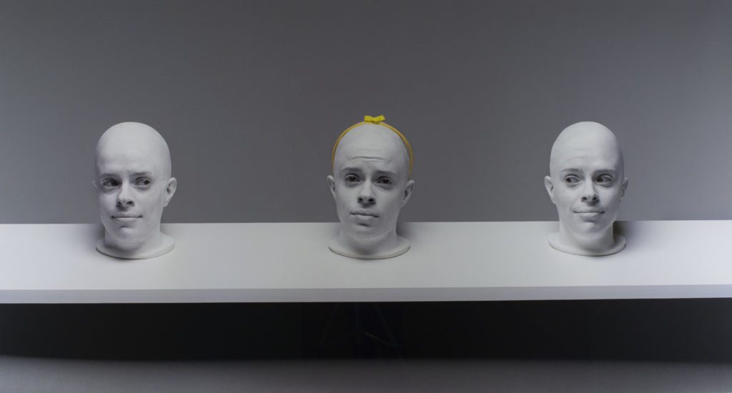 Photograph of three heads in white on a white shelf with a grey background. The center head has a yellow headband with a bow on its head.