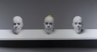 Photograph of three heads in white on a white shelf with a grey background. The center head has a yellow headband with a bow on its head.