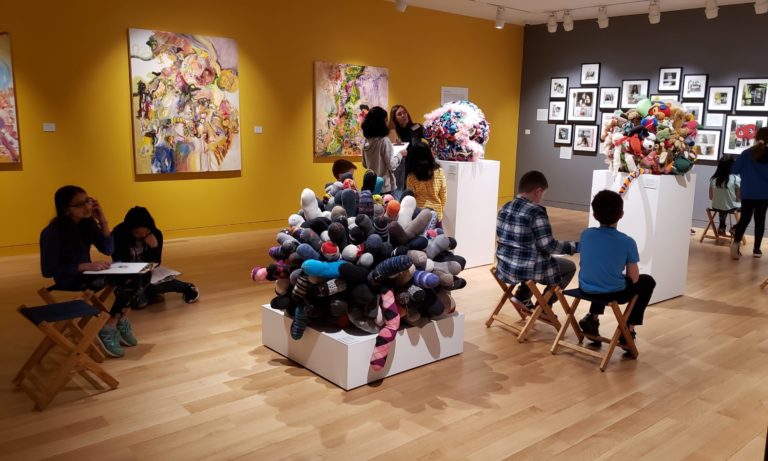 Photograph of a gallery room with yellow and grey walls, paintings on the yellow wall and photographs on the grey, with white pedestals holding balls made of various fabrics. Around this standing and sitting are young students drawing.