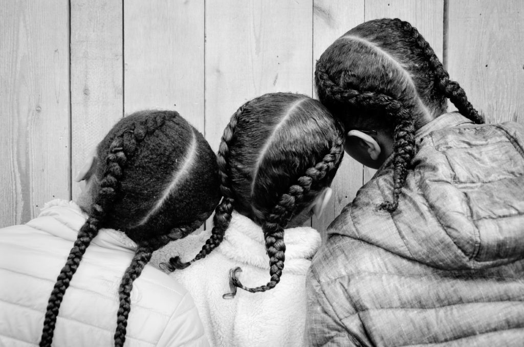 Black and white photograph of the back heads of three young girls, all with long braids on either side of their heads.