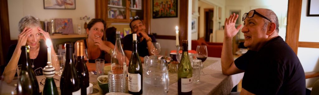 Long photograph of four people seated around a dining table covered in wine bottles, glasses, and candles, actively in some amusing debate.