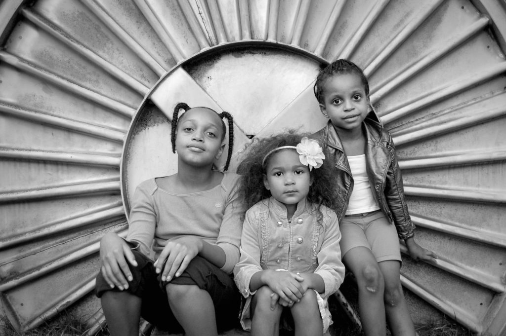 Black and white photograph of three young African American girls seated in a large round metal circle with rippled edges.
