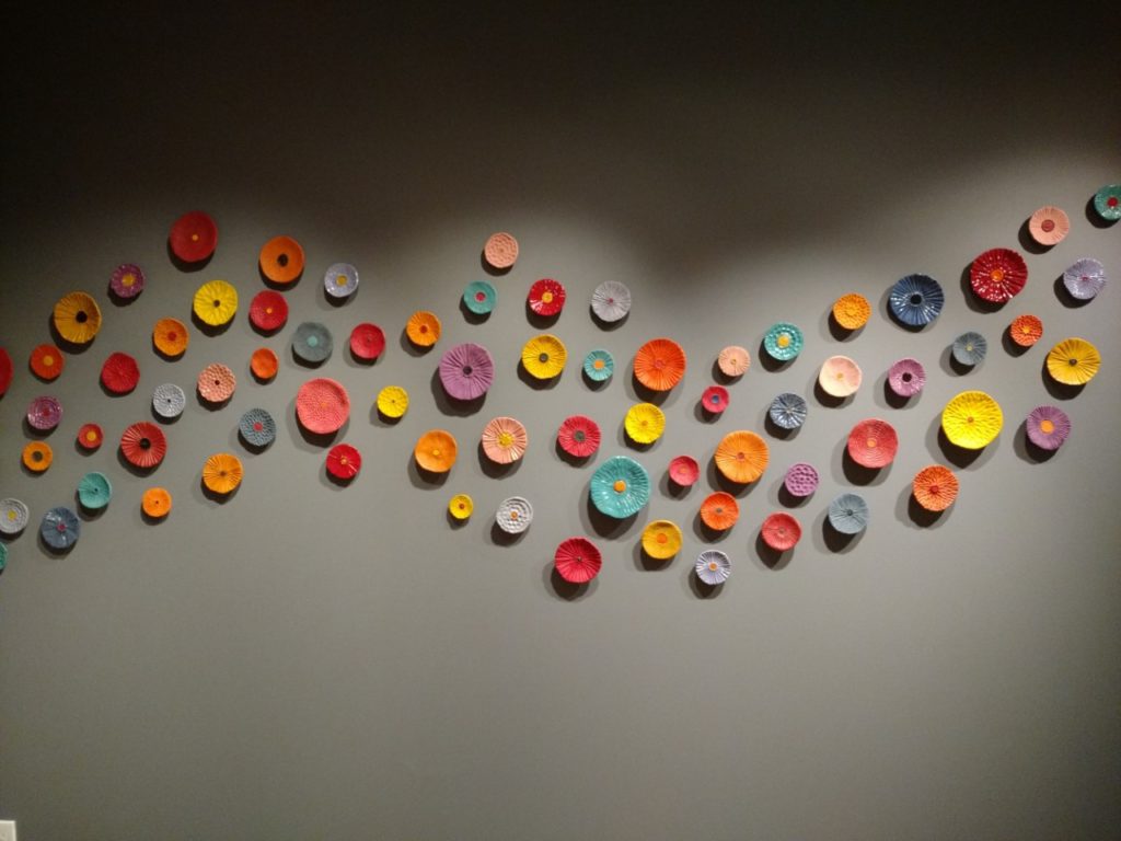 Photograph of a wall installation of dozens of ceramic round flowers in shades of orange, yellow, purple, and blue, all on a dark grey wall.