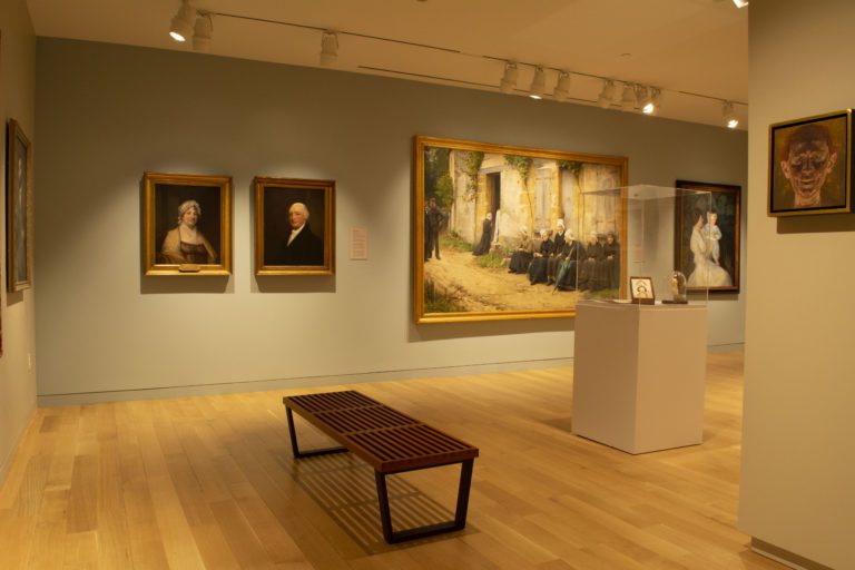 Photograph of a gallery room with light blue and grey walls and a bench in the center of the tan wood floor. There are several paintings hanging on all visible walls. A pedestal with cover is also visible with several small sculpture pieces.