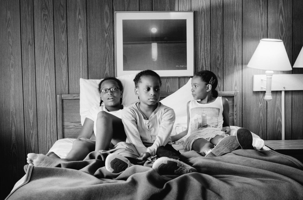 Black and white photograph of three African American girls in a hotel bed with a photograph hanging above them and a pair of wall lights to their right.