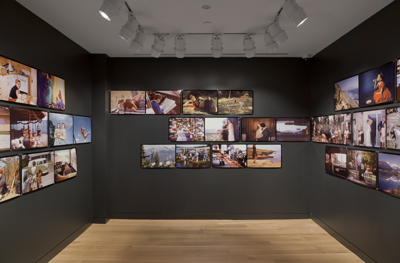 Photograph of a gallery room with dark grey walls and tan wood floor. There are large photographs in film strip style on the walls in rows of three.