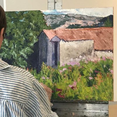 Photograph of a student painting, seen to the left, wearing a grey and white stripped shirt. They are standing at an easel painting a landscape scene with white washed building in the center with a red roof.