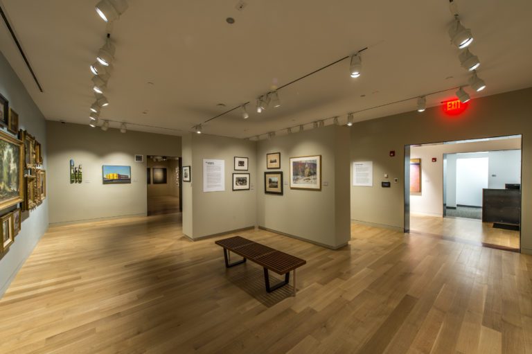 Photograph of a gallery room with light blue and tan walls and light wood floor with a bench. There are various pictures on all walls, including many 19th century works in gold frames on the left side hung close together at different levels.