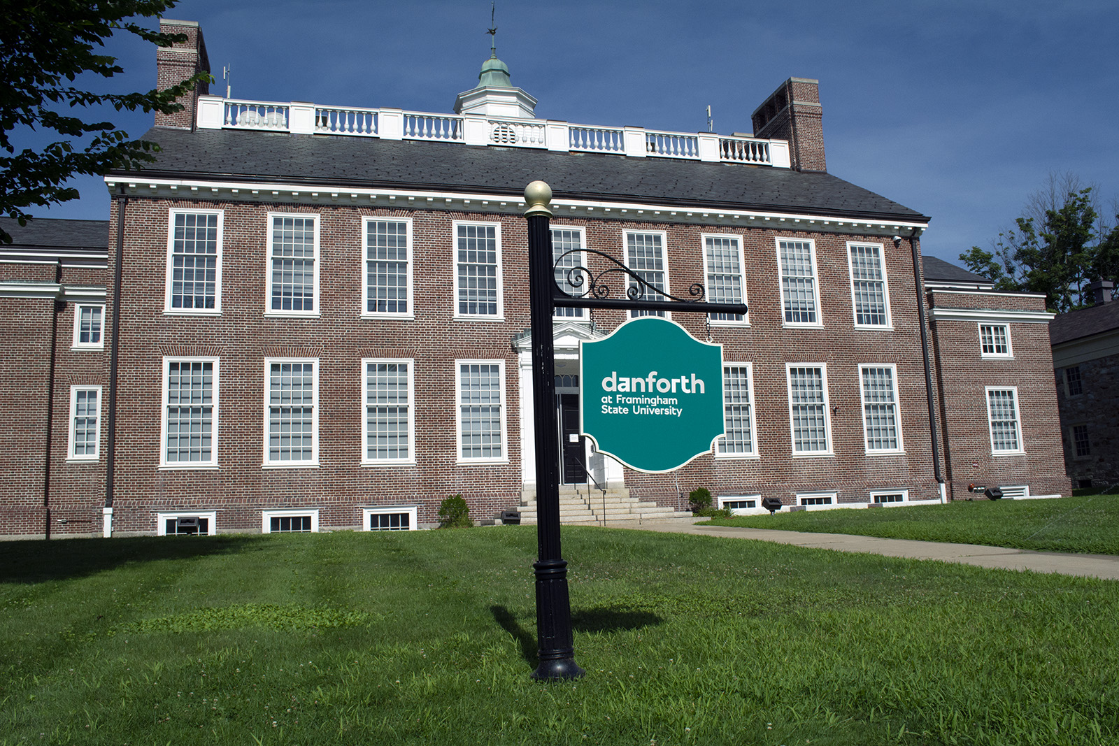 Photograph of a two story brick building at a slight angle. The windows and door are framed in white. The roof has tall brick fireplace in a rectangle on the far right, white fencing, and a cupola with a green roof in the center. There is a long green lawn with teal sign and a bright blue sky.