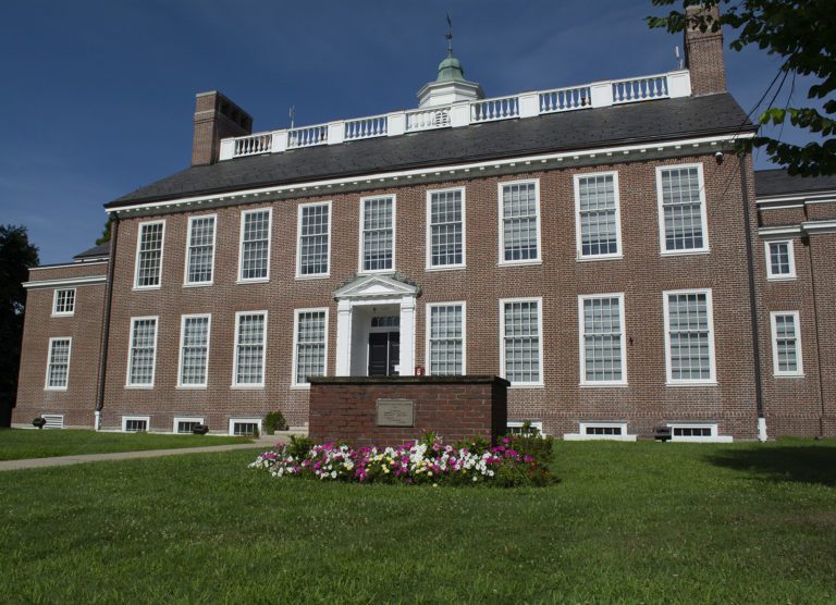 Photograph of a two story brick building at a slight angle. The windows and door are framed in white. The roof has tall brick fireplace in a rectangle on the far right, white fencing, and a cupola with a green roof in the center. There is a long green lawn and a bright blue sky.