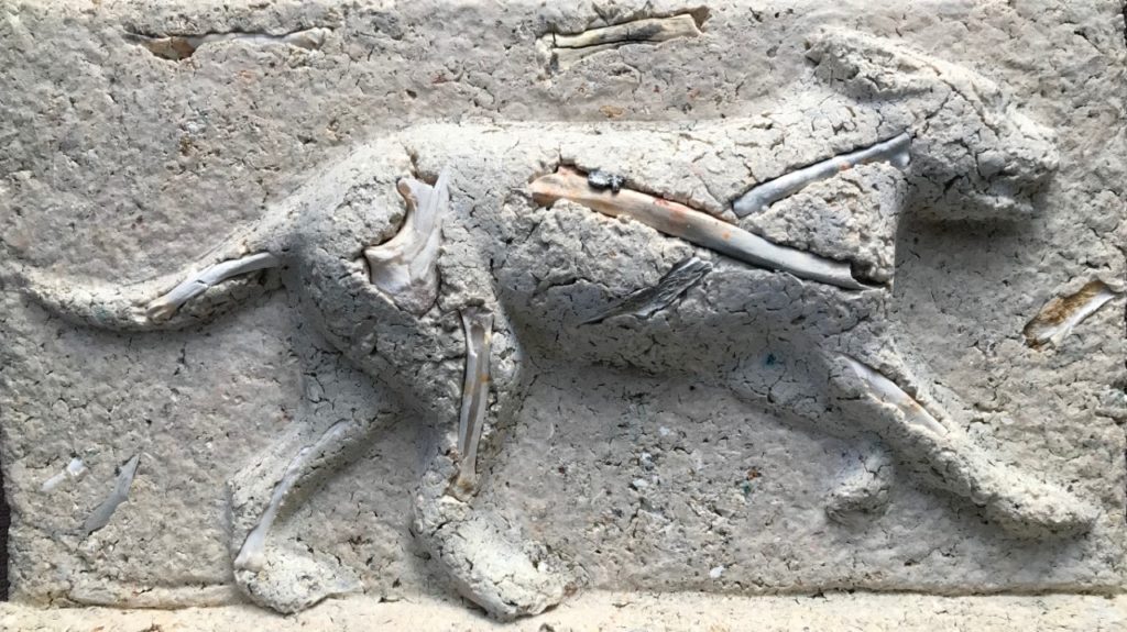 Sculpture in grey-white of a dog running to the right. There are bits of burnt bone visible throughout.