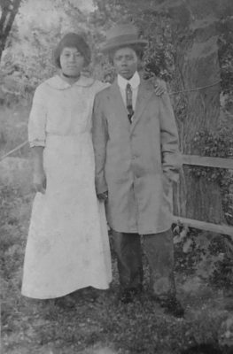 Old black and white photograph of an African American women couple, one dressed in a man's suit with a long coat and hat. The woman on the left is wearing a white dress. They are outside standing by a tree and wooden fence.