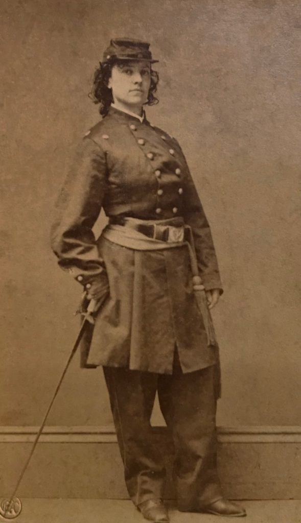 Old yellow tinted black and white photograph of a woman, standing, with curly hair. The woman is wearing a civil war uniform with a sword in her left hand.