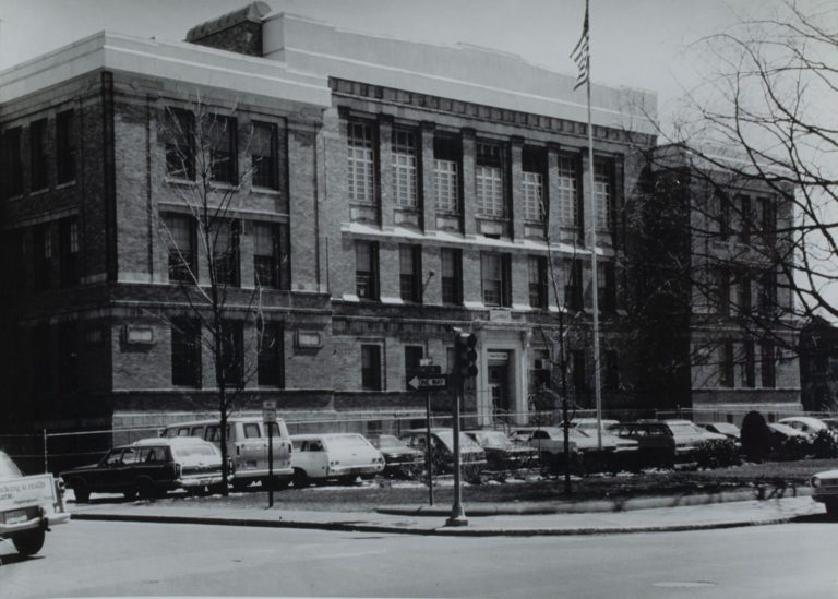 1970s black and white photograph of a three story building. There is a flag pole out front and a row of old 1960s and 1970s cars parking in front of the building.