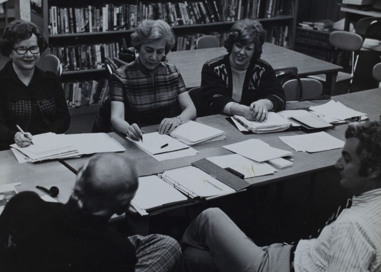 1970s black and white photograph of a group of people at a long table with many papers in front of them. The focus is on three women on the far side of the table, two men sit opposite seen from behind.