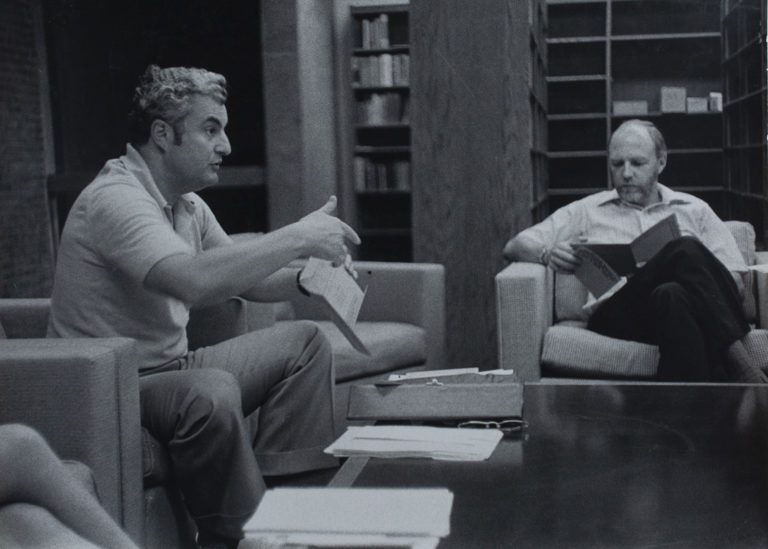 1970s black and white photograph of a two men seated in arm chairs. The man on the left is in active discussion with someone off view.