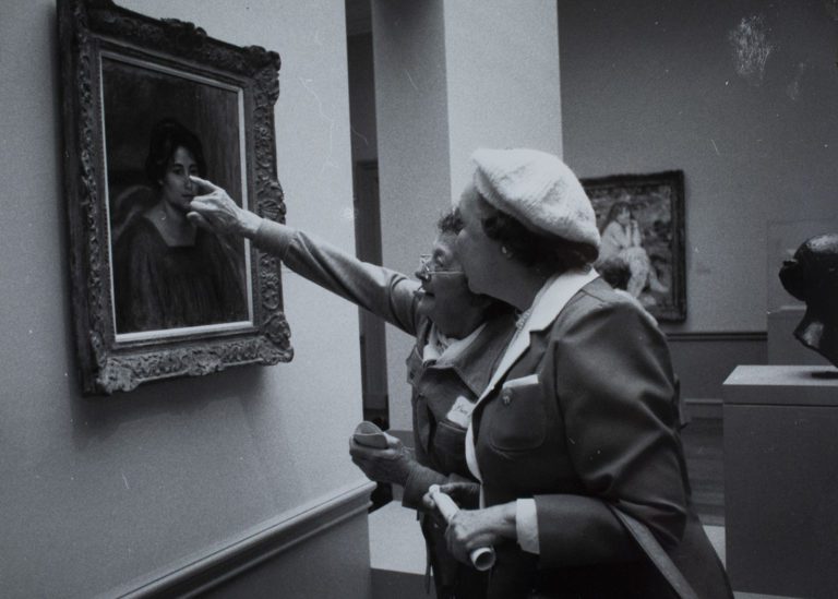 1970s black and white photograph of two older women looking at a 20th century painting hanging on a wall. One woman is reaching towards the painting, pointing out a detail on the figure's face.