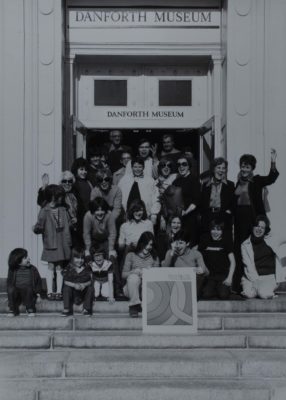 Black and white photograph from 1975 of a large group of people on steps leading into a doorway to a building. There is a poster being held on the steps by two kids.