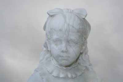Photograph of a girl in marble with pulled back long hair with a bow and a ruffled high collard top. A shadow leaf pattern has been imposed over her face. The sky is grey.